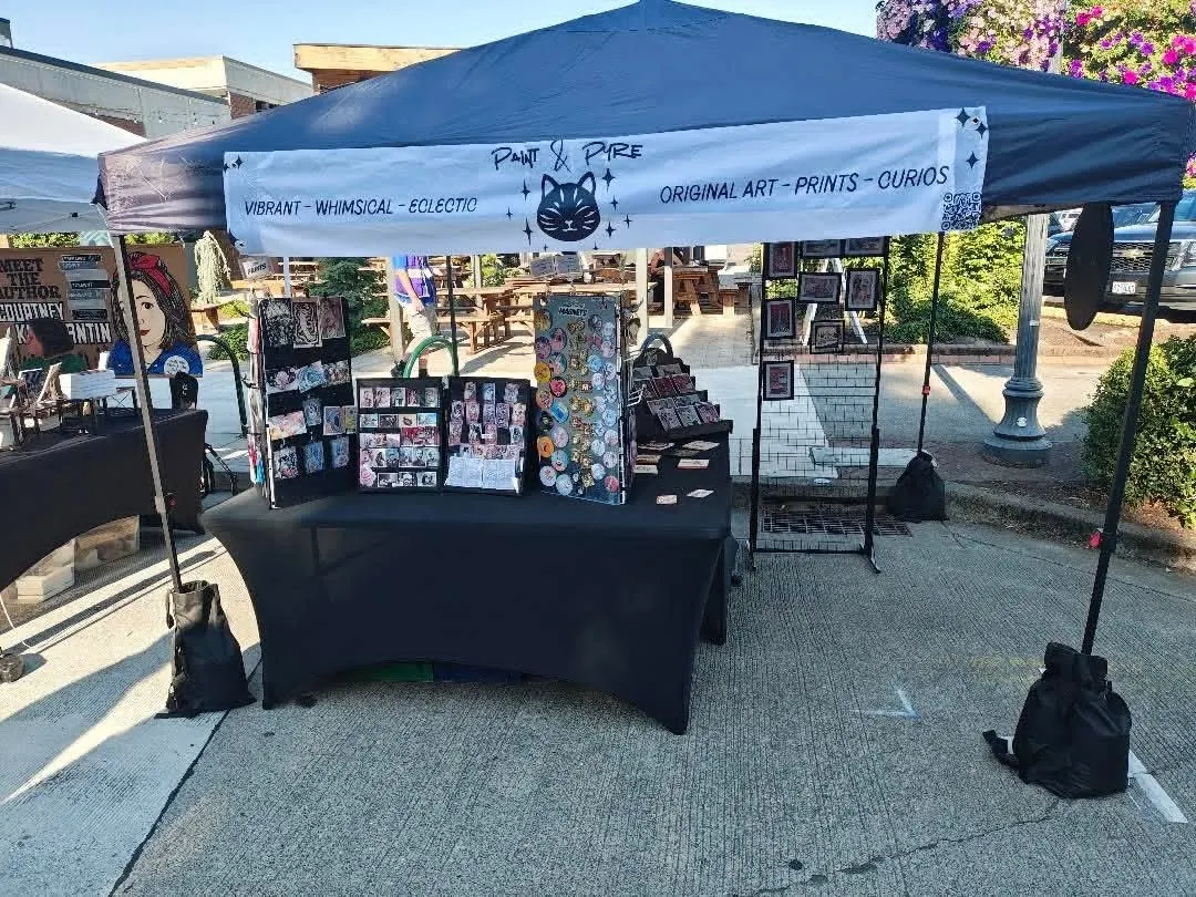 The Paint & Pyre stall set up in daylight, her printed banner across the front and racks of prints and magnets on the table
